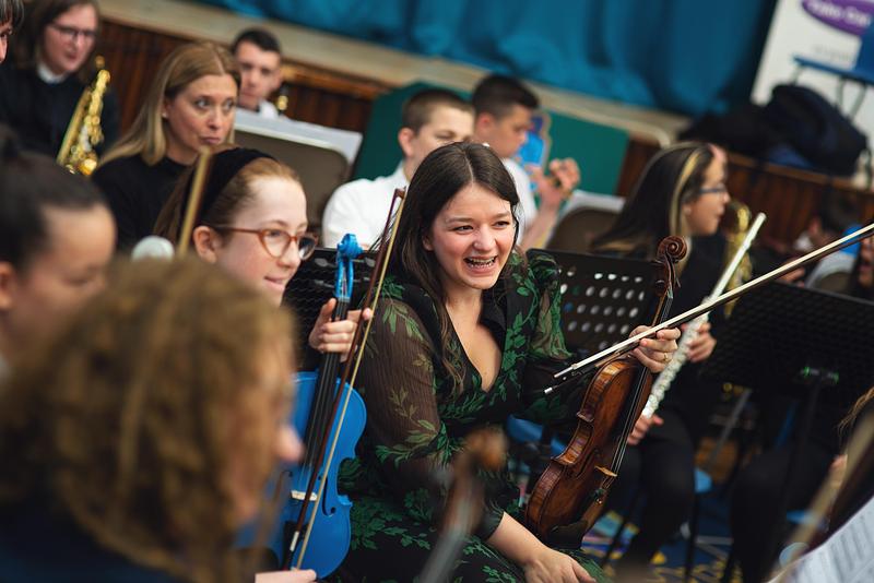 A member of the Royal Scottish National Orchestra (RSNO) and a pupil, both holding violins, smile in the middle of a school orchestra